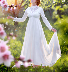 young beautiful red-haired Caucasian girl in a white dress enjoys flowering trees in the spring park