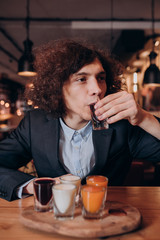 young curly-haired guy in a black suit drinking a liqueur in the bar