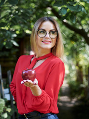 Young blond woman, wearing eyeglasses and red blouse, holding red claret apple in her hand....
