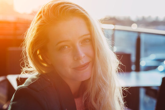 candid portrait of pretty smiling young woman with blonde hair wearing jacket enjoying beautiful sunset at city cafe, sunny day