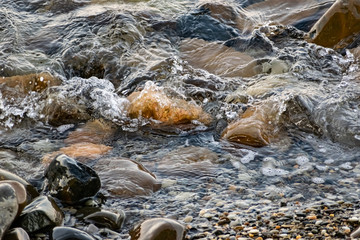Large and smooth stones in the coastal waves of salty sea water. The coast of the black sea foams and roars, beating against a huge pebble, illuminated by sunlight