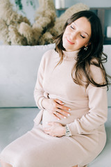 portrait of a young beautiful pregnant woman in a beige dress sitting on a gray sofa in a cozy Studio