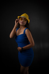 Fototapeta premium fashion portrait of an young and attractive Indian Bengali brunette girl with blue western dress, yellow cap and sunglass in front of a black studio background. Indian fashion portrait and lifestyle.