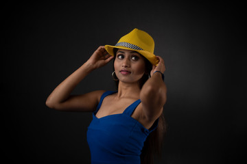 fashion portrait of an young and attractive Indian Bengali brunette girl with blue western dress, yellow cap and sunglass in front of a black studio background. Indian fashion portrait and lifestyle.
