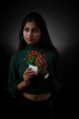 Fashion portrait of young and attractive Indian Bengali brunette girl with green cropped top and skirt holding cherry tree in front of black studio background. Indian fashion portrait and lifestyle.