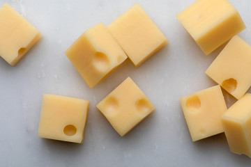 Portions (cubes, dice) of Emmental Swiss cheese with scratcher. Texture of holes and alveoli. On white marble background.