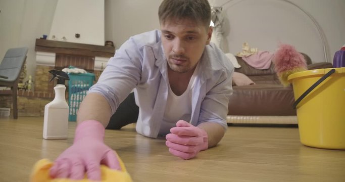 Close-up of young Caucasian man in protective gloves rubbing floor with cleansing agent. Tidy handsome brunette housekeeper cleaning parquet indoors. Household, domestic duty. Cinema 4k ProRes HQ.