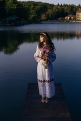 Fototapeta premium the bride in a chic embroidered dress stands on the pier against the background of the lake. girl holding a wedding bouquet.