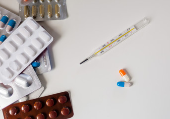 A large number of drugs on a white background next to a thermometer and two capsules of orange and blue