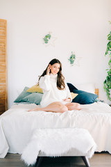 portrait of a young happy pregnant woman in a white warm sweater sitting on the bed and holding her stomach