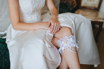 The bride sits on a sofa in a white dress with a garter on her leg. Morning preparation for the wedding. Photography, concept.