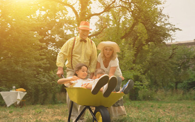 The Elderly Couple Is Pushing Their Grandchildren In A Wheelbarrow.All Members Of The Family Are Very Happy.Concept Of Active Vacation
