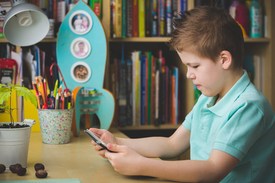 Primary School Student Is Sitting At Home At The Table And Working On A Tablet Pc. The Child Looks At The Tablet Developing Programs. Early Development And Learning. Toning