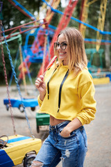 Young blond woman, wearing yellow hoody and blue jeans, holding colorful sweet tasty lollypop in...