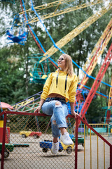 Young blond woman, wearing yellow hoody and blue jeans, spending time in amusement theme park in summer. Three-quarter portrait of pretty girl, sitting on railing in front of colorful ferris wheel.