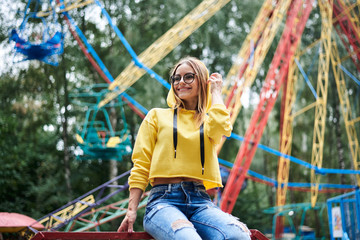 Young blond woman, wearing yellow hoody and blue jeans, spending time in amusement theme park in summer. Three-quarter portrait of girl, sitting on railing in front of colorful ferris wheel, smiling.