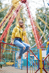 Young blond woman, wearing yellow hoody and blue jeans, spending time in amusement theme park in summer. Three-quarter portrait of pretty girl, sitting on railing in front of colorful ferris wheel.