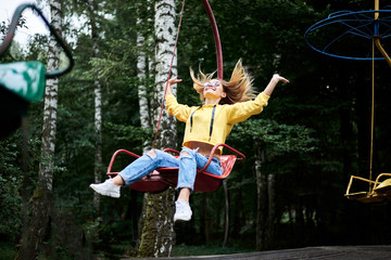 Young blond woman, wearing yellow hoody and blue jeans, riding on carrousel, smiling, laughing in park with green trees in summer. Entertainment in amusement park. Sunday leisure time.