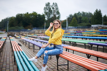 Young blond woman, wearing yellow hoody, blue jeans and eyeglasses, sitting on colorful benche in city urban park in summer. Portrait of pretty girl, relaxing resting on sunday afternoon. Leisure time