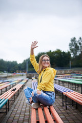 Young blond woman, wearing yellow hoody, blue jeans and eyeglasses, sitting on colorful benche in city urban park in summer. Portrait of pretty girl, relaxing resting on sunday afternoon. Leisure time