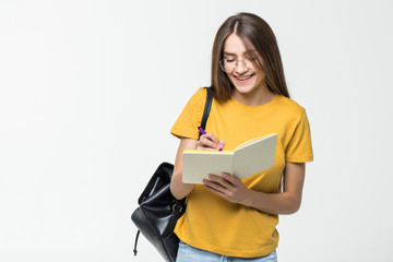 Portrait of a smiling casual girl student with backpack writing in a notepad while standing with books isolated over white background