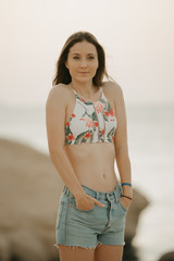 A pretty model with long hair posing with hands into the pockets of the denim shorts on the rocky shore of the red sea in a swim top on the sunrise