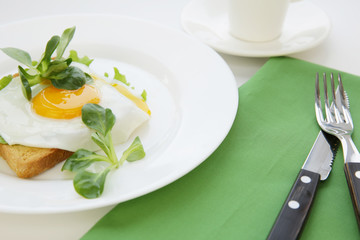 fried egg with a yellow yolk on bread toast and green salad on a white plate and a cup of coffee or tea. Table setting with green napkin, fork and knife on a white table. Tasty and healthy breakfast.