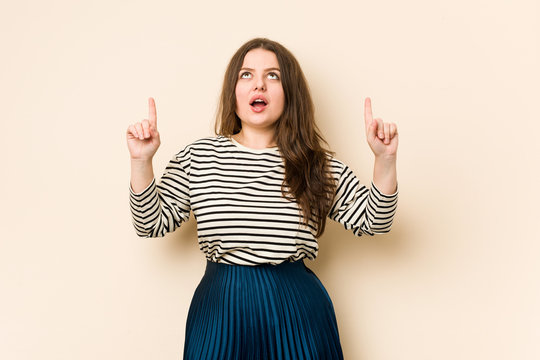Young curvy woman pointing upside with opened mouth.
