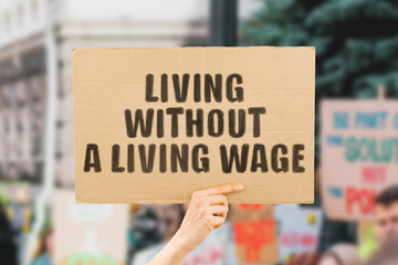 The phrase " Living without a living wage " on a banner in men's hand. Human holds a cardboard with an inscription. Financial. Poorness. Money. Earnings. Revenue. Life. Social security. Economy