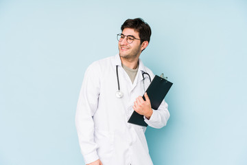Young doctor man isolated on blue background looks aside smiling, cheerful and pleasant.