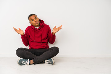 Young latin man sitting on the floor isolated doubting and shrugging shoulders in questioning gesture.