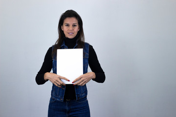 Portrait of attractive caucasian middle age woman holding white empty paper, isolated on gray background studio shot, black sweater, denim jacket, jeans. Place for your text in copy space.