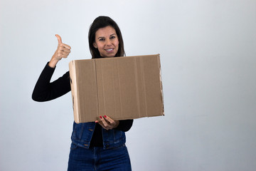 Portrait of attractive caucasian middle age woman showing thumb up gesture with a cardboard box, isolated on gray background studio shot, black sweater, denim jacket, jeans, dark air. Place for your t