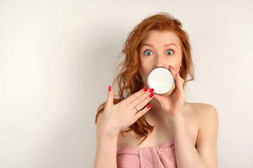 Redhead girl with clean skin looking at the camera with eyes wide open and covering mouth with a jar of cream, putting hand to her