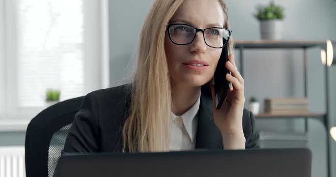 Mature blonde manager in black suit and eyewear having phone conference at modern office while working on laptop. Businesswoman talking on smartphone at working place.