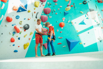 Father helping his teenage son and checking climbing harness near the indoor climbing wall. Boy...