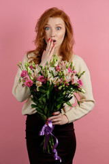 Beautiful redhead girl with a bouquet of flowers on a pink background shockingly covering mouth with hand, looking at the camera
