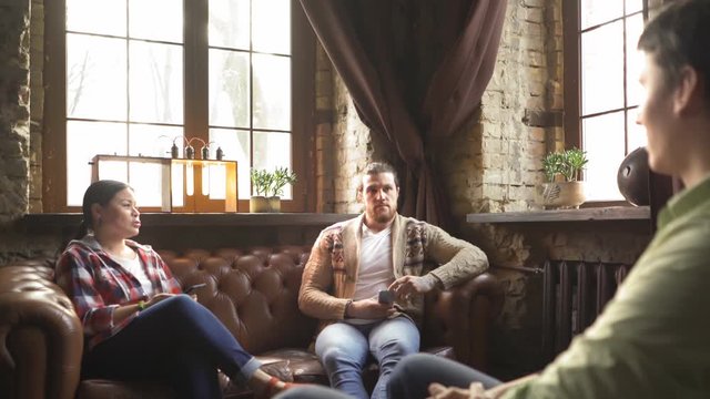 Multiethnical group of three people men and women sitting on a counch in a modern office. Mid aged asian woman sitting with freelancers coworkers on a leather sofa. Students meeting with their curator