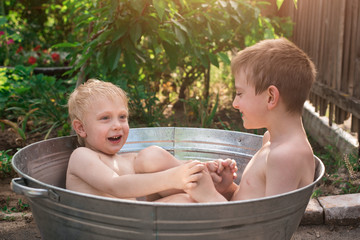 Two boys sitting in metal basin full of water and play. Children bathe in basin outside