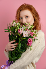 Beautiful redhead girl presses a bouquet to her face and looks cheerfully at the camera through the flowers, on a pink background