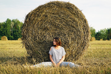Naklejka na ściany i meble A young girl is sitting near a round stack of straw in a mown field of rye.