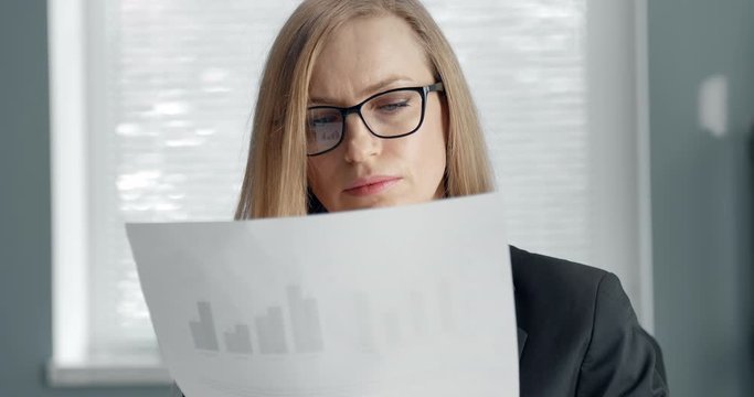 Portrait of mature business woman with blond hair carefully reading important document while sitting at own office. Beautiful lady in formal clothing and eyeglasses checking annual report at work