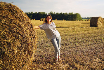 Naklejka na ściany i meble A young girl stands near a round stack of straw on a mown field of rye.