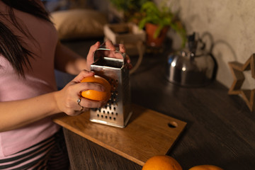 Young woman in pajamas rubbing fruit on grater. Women's hands doing zest from orange in kitchen.