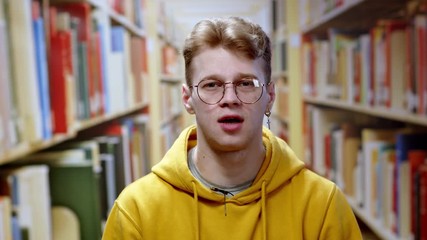 Young speaker handsome student talking on camera job interview online course reportage looking at webcam near bookshelves staying in the library.