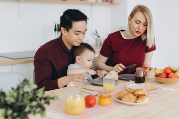 Happy multicultural family. Asian dad and his caucasian blonde wife have breakfast with their beautiful son in the kitchen.