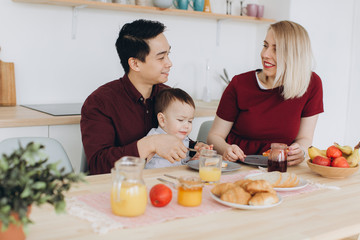 Obraz na płótnie Canvas Happy multicultural family. Asian dad and his caucasian blonde wife have breakfast with their beautiful son in the kitchen.