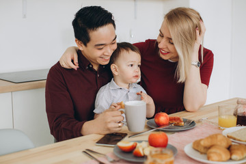 Happy multicultural family. Asian dad and his caucasian blonde wife have breakfast with their beautiful son in the kitchen.