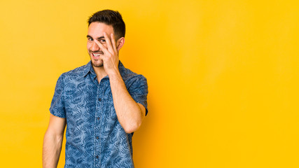 Young caucasian man isolated on yellow bakground blink at the camera through fingers, embarrassed covering face.