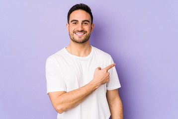 Young caucasian man isolated on purple background smiling and pointing aside, showing something at...